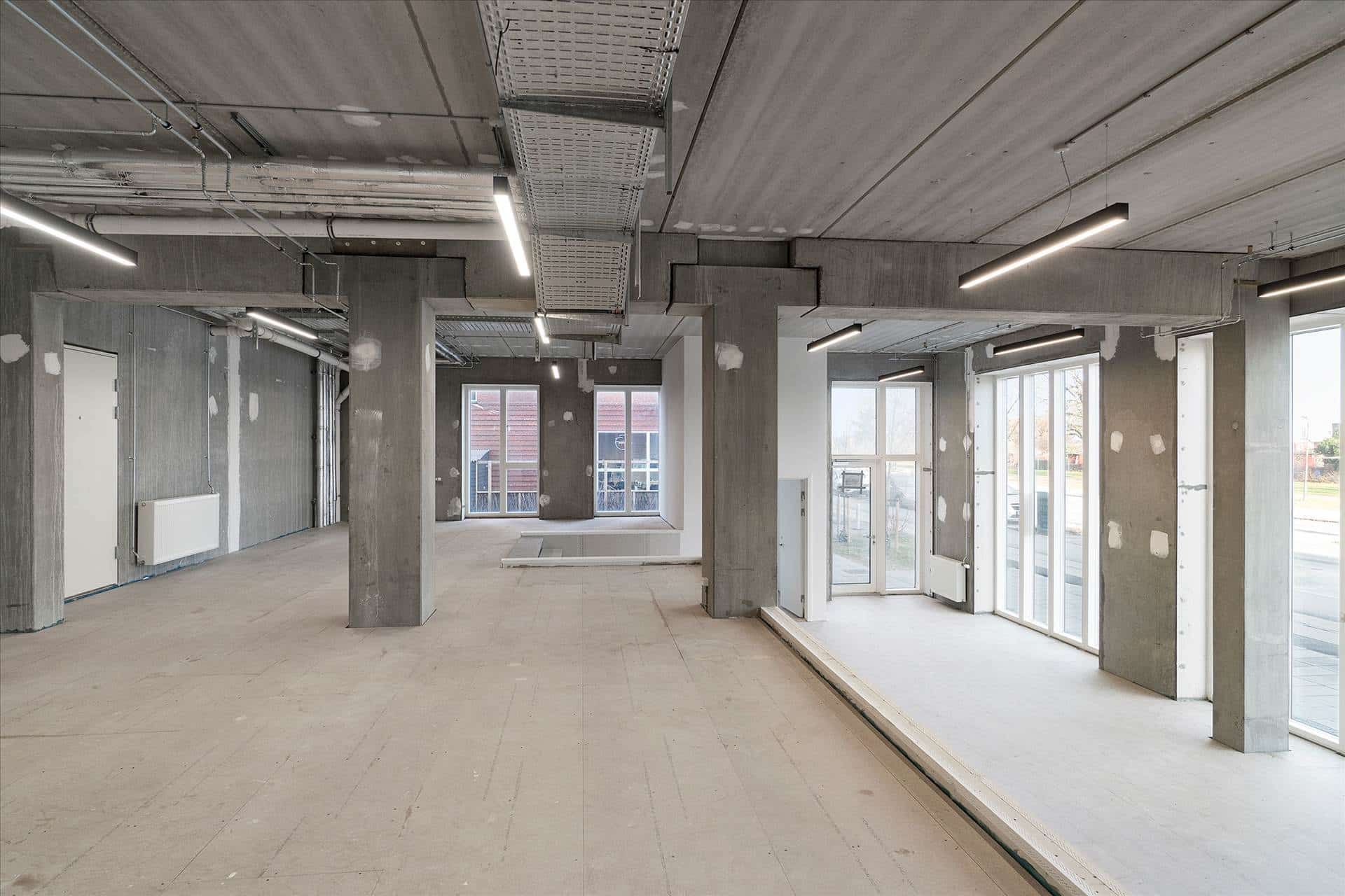 Inde i en tom værkstedbygning med højt loft, slanke betonpelare, hvide gulve og store vinduer. Der er lysekroner og hængende