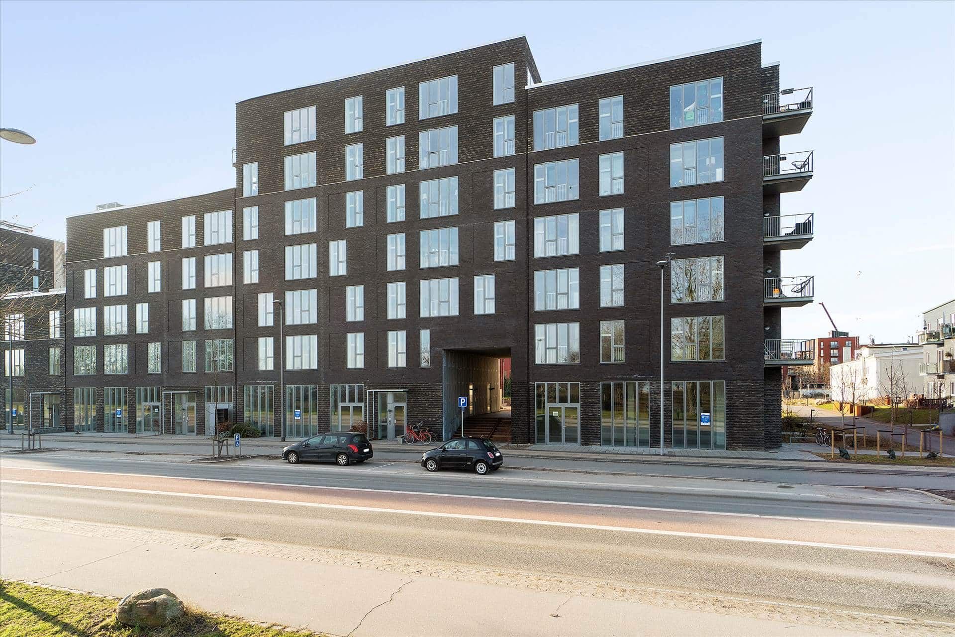 Moderne og høj bygning med mange vinduer og store balkoner. To biler parkeret foran bygningen.