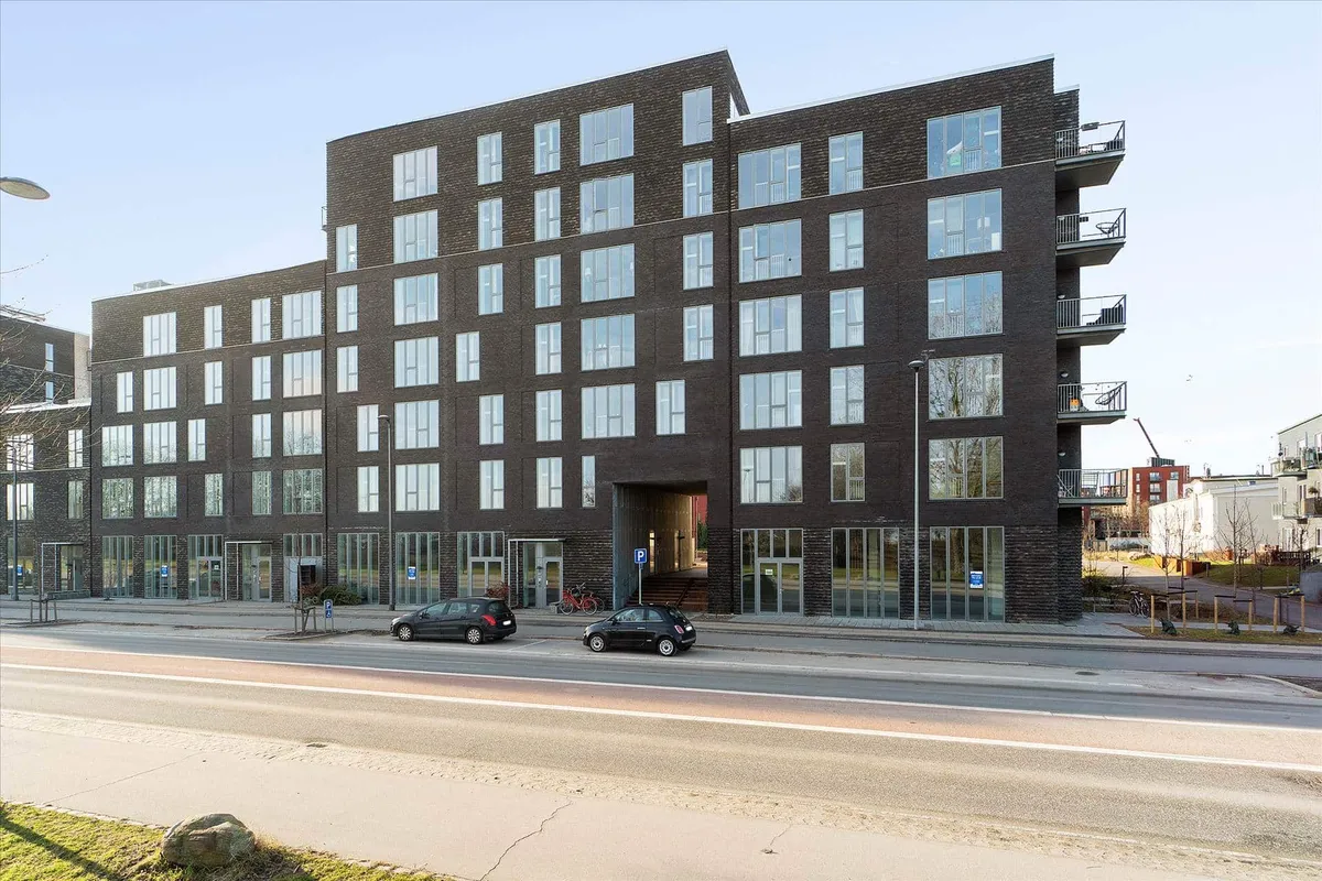 Moderne og høj bygning med mange vinduer og store balkoner. To biler parkeret foran bygningen.