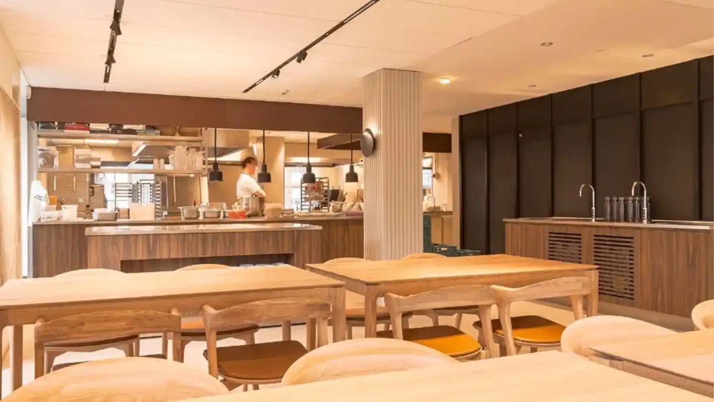 En moderne køkkenlokaler med lyse træborde og stole, samt en person ved en stolpe med loftslamper