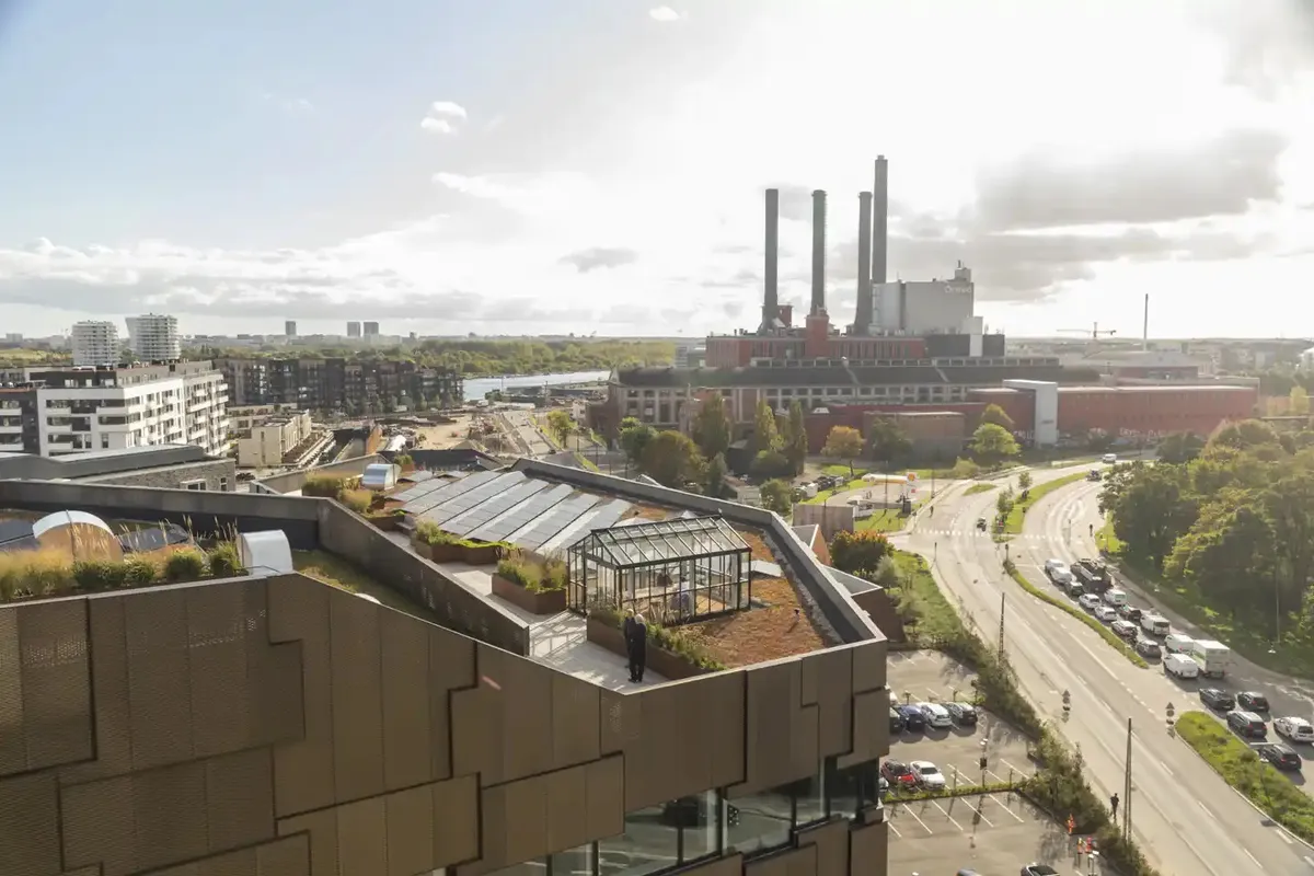 En bygning med grøn tagterrasse og solceller i byens centrum med flere biler parkeret og en stor vej med trafik