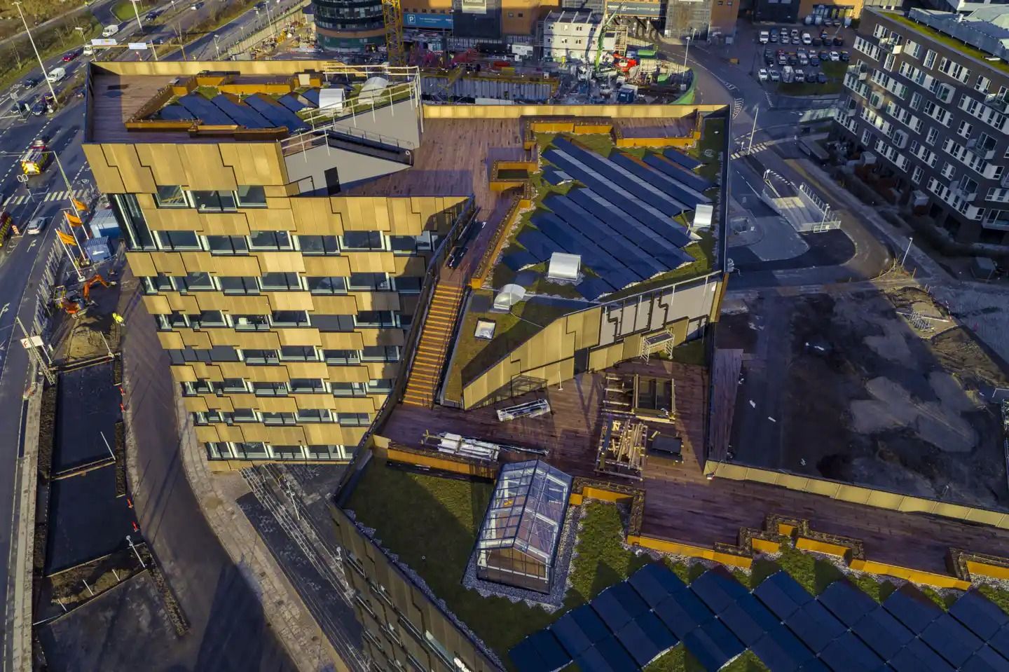 Bygningen med solceller og terrasse er under opførelse med snedækkede tag. Mange biler parkeret og bygninger i baggrunden