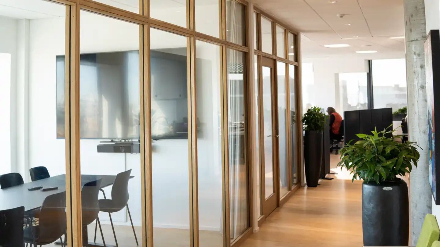 Kontorrum med glas og træpaneler, møbler og planter samt en person i orange vest.