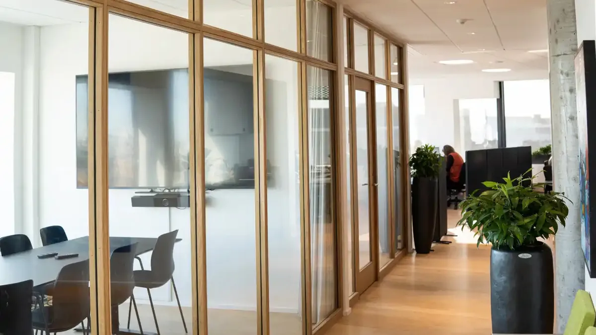 Kontorrum med glas og træpaneler, møbler og planter samt en person i orange vest.