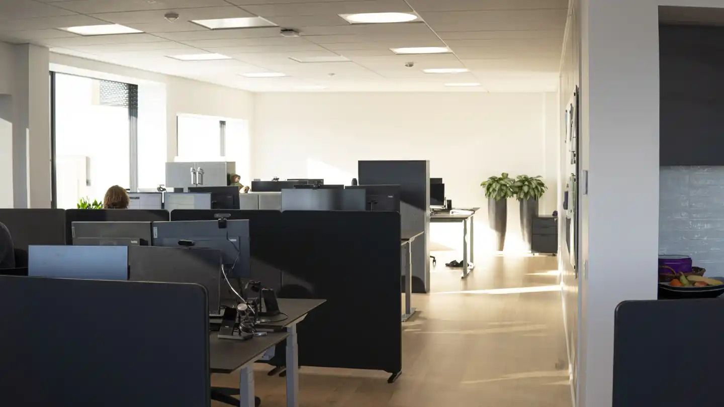 Et moderne kontor med en hvid væg og højt loft med lysepunkter og vinduer med gardiner. Der er flere skriveborde med