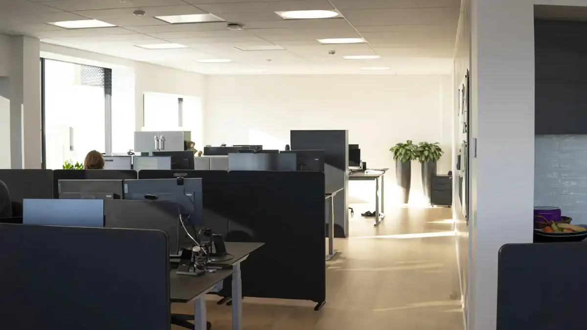 Et moderne kontor med en hvid væg og højt loft med lysepunkter og vinduer med gardiner. Der er flere skriveborde med