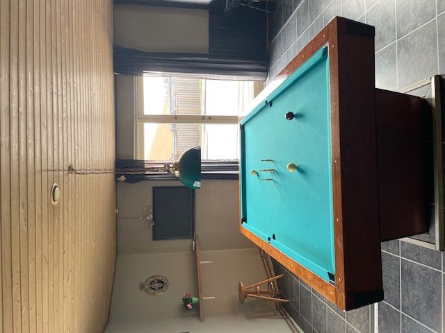 Poolbordet er monteret på væggen med en stol til siden og en grøn lampe. Væggene er klædt med træ og mørke gardiner. Et