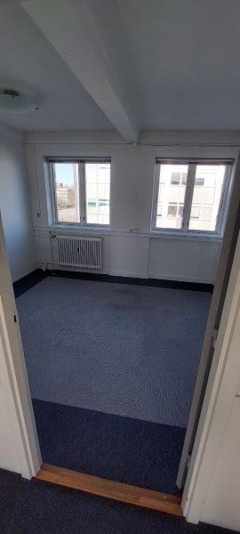 En tom kontorrum med to vinduer, hvor der er hængt radiatorer på væggen og der er blå gulvbelægning