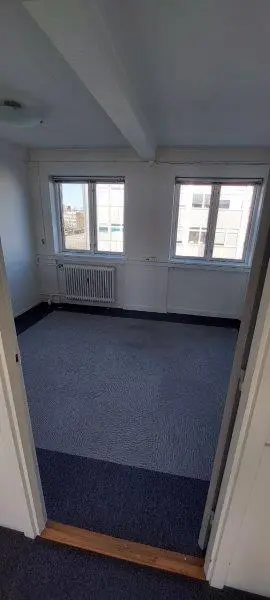 En tom kontorrum med to vinduer, hvor der er hængt radiatorer på væggen og der er blå gulvbelægning