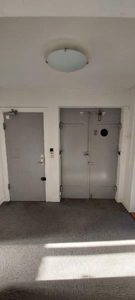 To høje døre i lysegråt, sammen med en hvid væg og et rundt loftslys. Gulvet er dækket med gråt gulvtæppe og solstråler