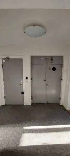 To høje døre i lysegråt, sammen med en hvid væg og et rundt loftslys. Gulvet er dækket med gråt gulvtæppe og solstråler