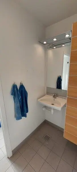 Et lille badeværelse med en hvid væg og to hængede blå håndklæder samt en spejl og et hvidt vasketøjsskab med en håndtag