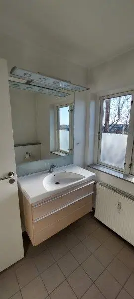 En moderne badeværelse med et stort spejl og en hvid vask samt en radiator under vinduet