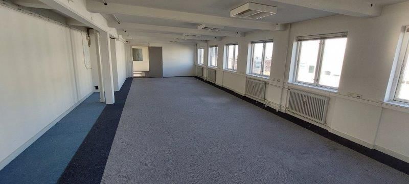 En tom og lys kontorlokal med hvide vægge og højt gulv med blåt gulvtæppe og flere vinduer med lyse gardiner