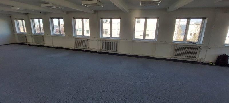 Rummeligt kontor med højt til loftet og store vinduer, hvor man ser ud på bygninger og grønne træer