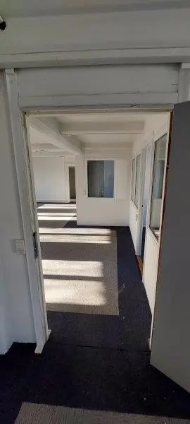 Et rummeligt kontor med hvide vægge og lofter, hvor sollys strømmer gennem de åbne døre og vinduer. Gulvet er dækket med en