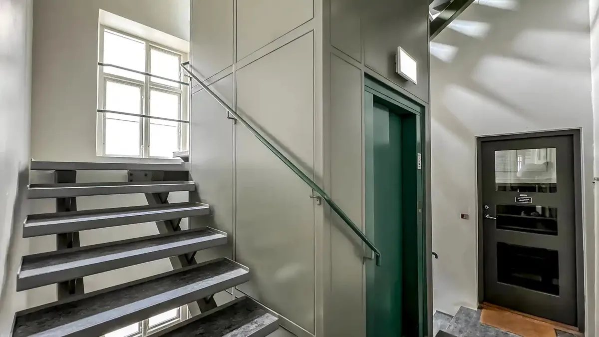 En indre trappe med skrappe grå trappetrin og grønne elevatordøre med metalhåndtag og glasvinduer i en minimalistisk