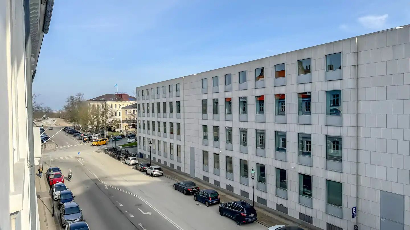 En bred gade med parkerede biler, en bygning med mange vinduer og en cykelstel på siden og busser og biler i baggrunden