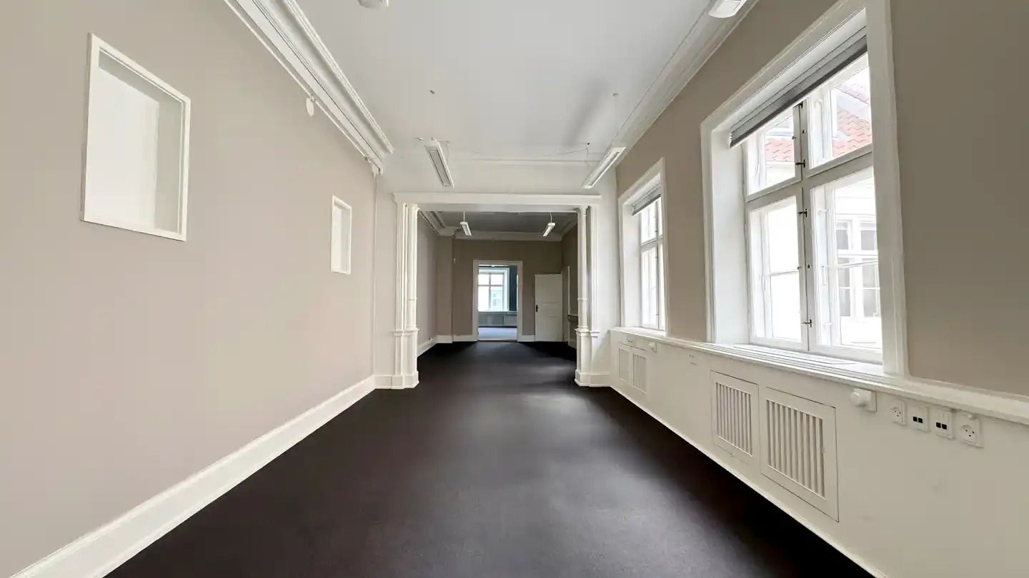 Et tomt og elegant kontor med hvide vægge og sort gulv, høje vinduer, lysekroner og en dør i midten