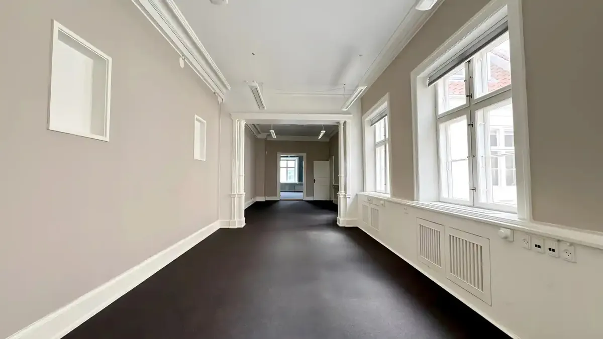 Et tomt og elegant kontor med hvide vægge og sort gulv, høje vinduer, lysekroner og en dør i midten