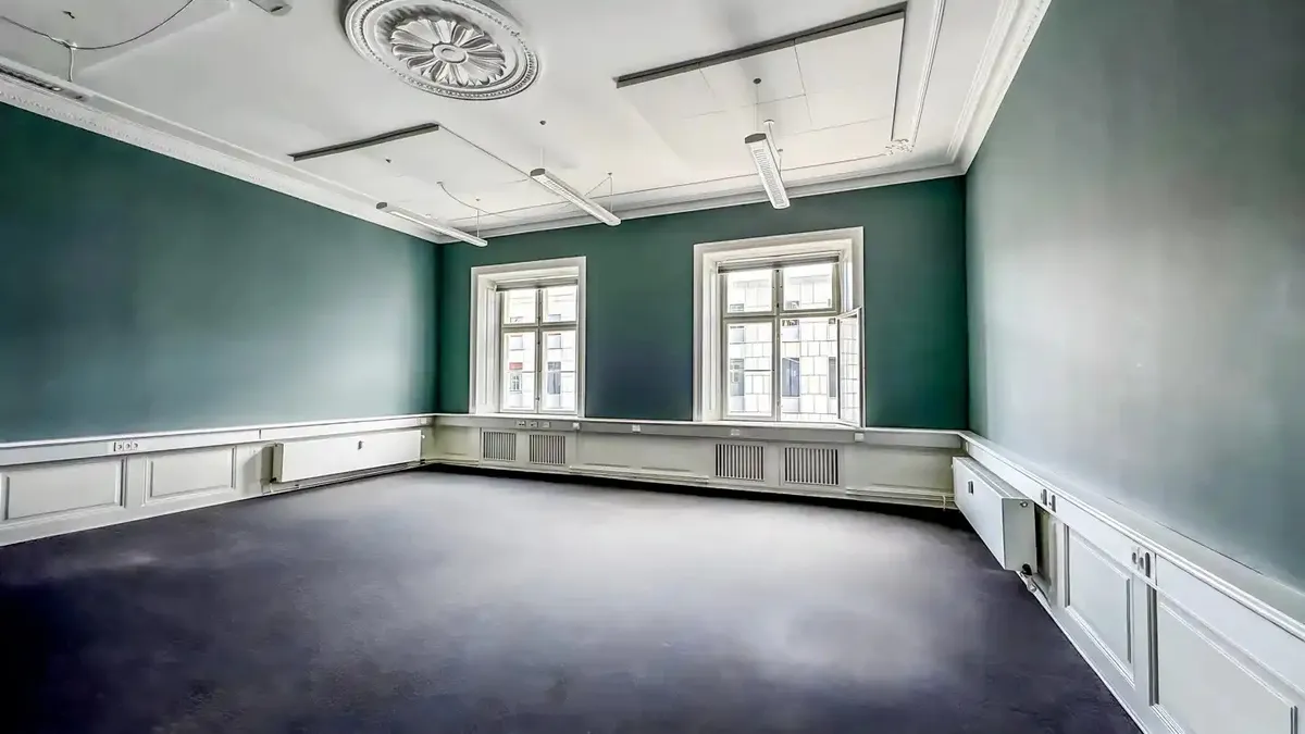 Kontorrum med højt til loftet, grønne vægge, hvide sokkelborde og to store vinduer med udsigt til bygningerne i baggrunden