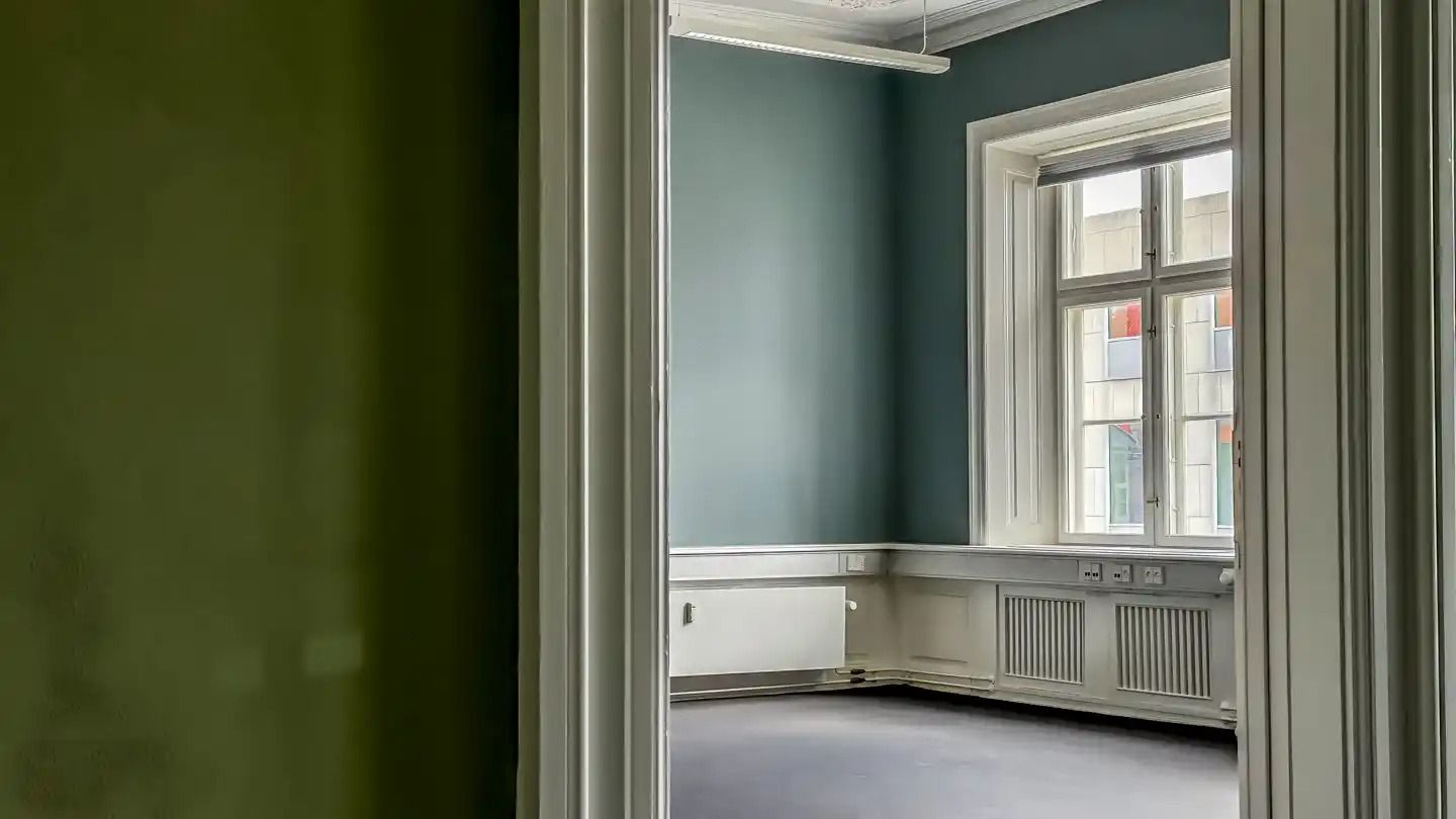 Et tomt kontorlokal med to vinduer og et radiatorpanel i væggen mod højre samt et grønt vægmaleri mod venstre