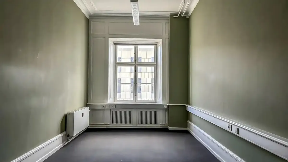 Kontorrum med grønne vægge, hvide paneler og en stor vinduesflade med udsigt til gaden udenfor, hvor en bygning er synlig