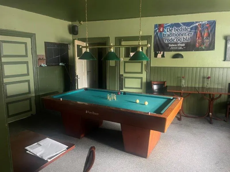 Billiardbord med poolkugler og kegler placeret i et værelse med grønne vægge og to træborde med glas og blomster.