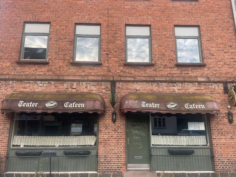 En lejlighedsbygning med to indgange til Café Teater, hvor det ene vinduesruder er mørke, og den anden har hvide gardiner,