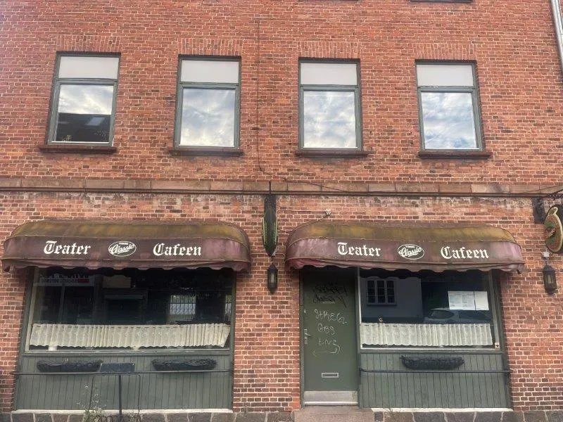 En lejlighedsbygning med to indgange til Café Teater, hvor det ene vinduesruder er mørke, og den anden har hvide gardiner,