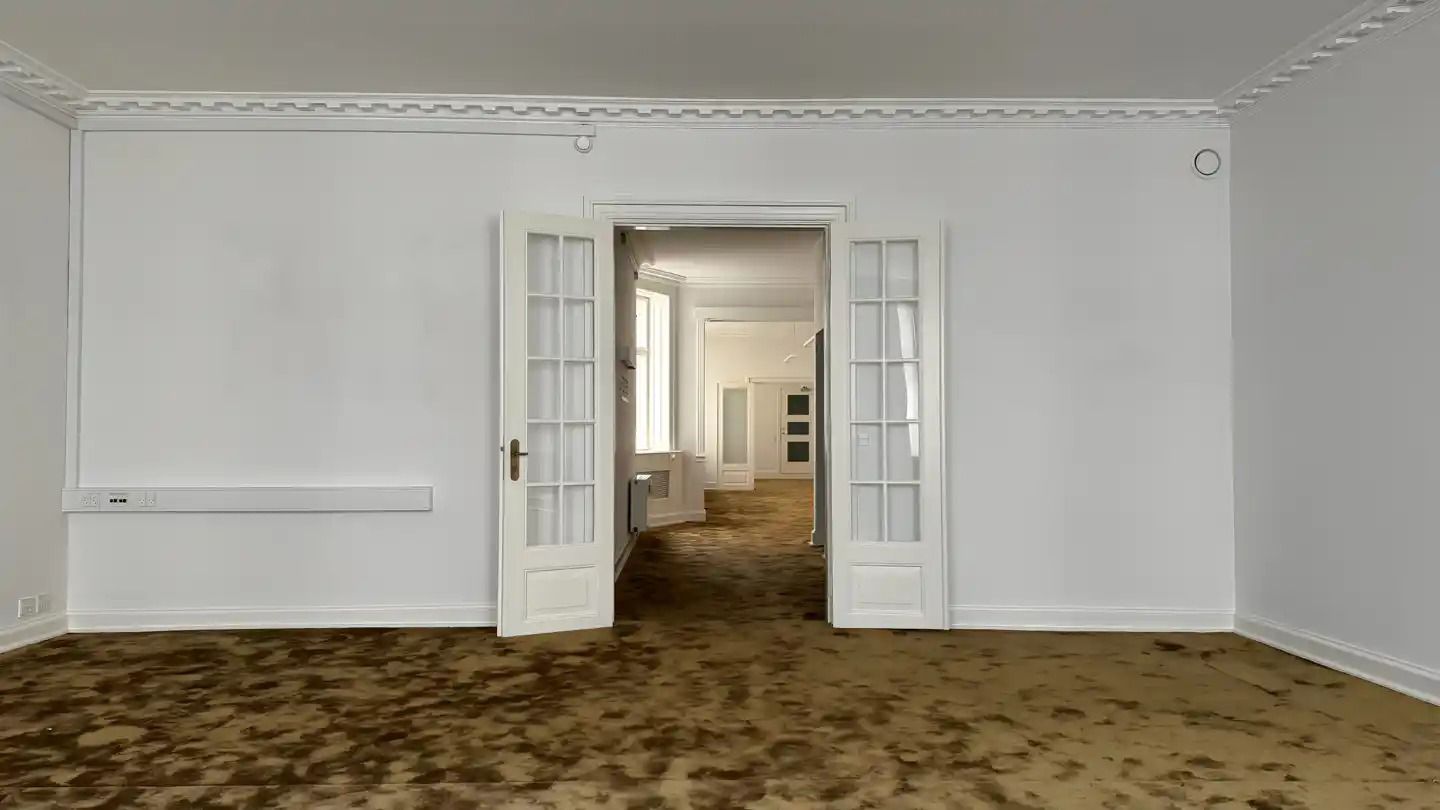 Moderne, lysfyldt stue med lysegul gulvbelægning og hvide vægge. Dørene er åbne og viser et rum med gulvbelægning og hvide