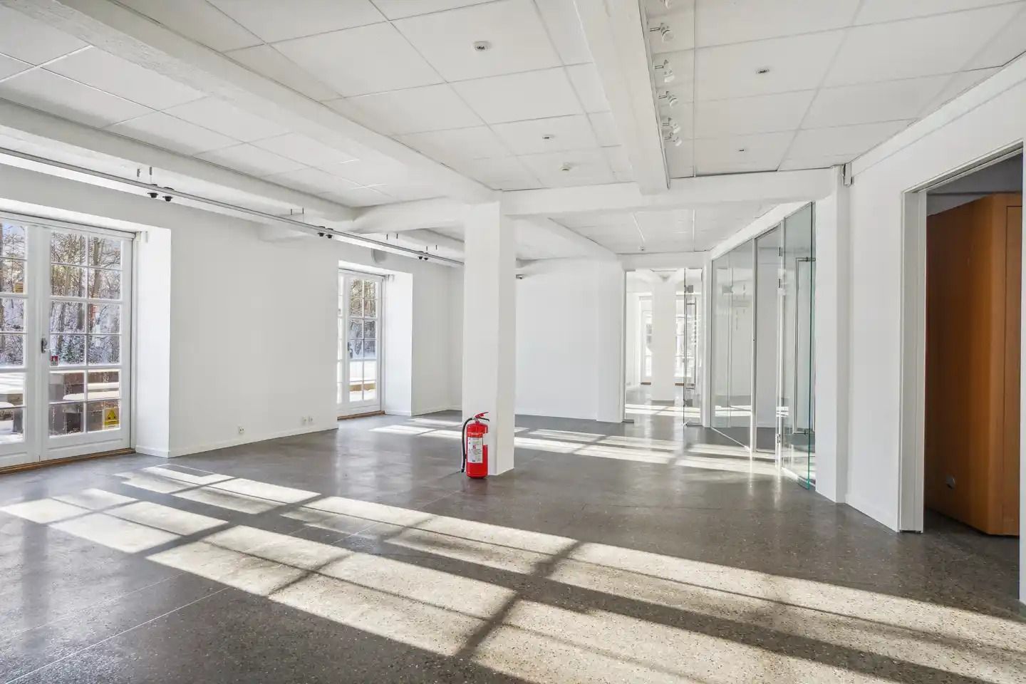 Minimalistisk kontor med hvidt loft, gulv og vægge, glasvinduer, rødt brandslukker, søjler og smukke skygger på gulvet fra