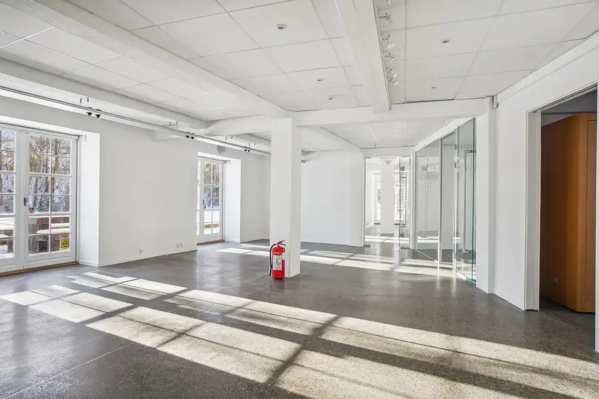 Minimalistisk kontor med hvidt loft, gulv og vægge, glasvinduer, rødt brandslukker, søjler og smukke skygger på gulvet fra