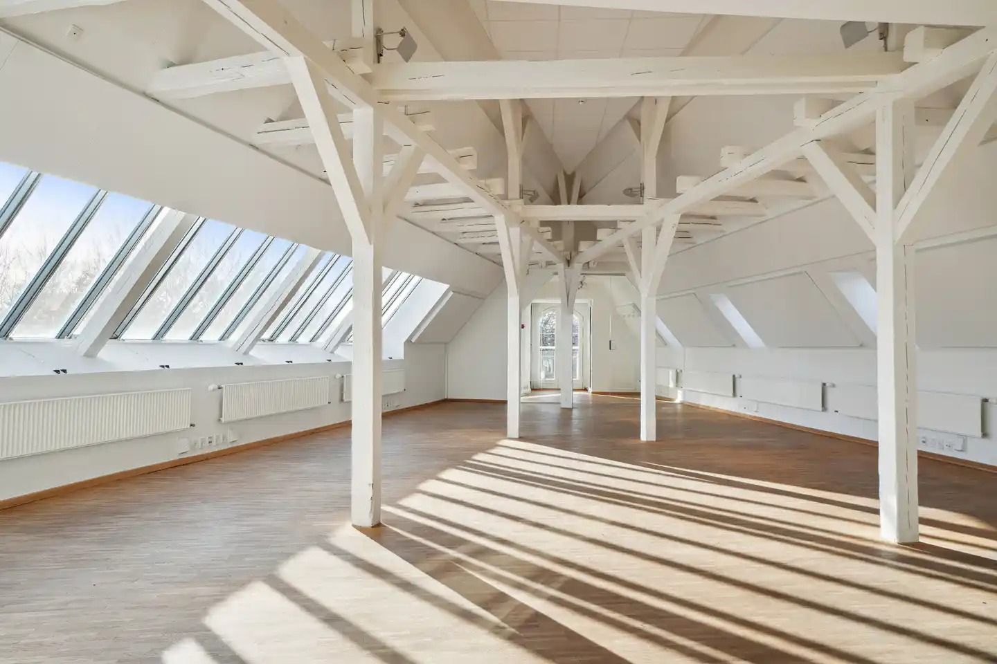 Lysfyldt loftsrum med hvide vægge og trægulv samt høje vinduer og lyse stråler