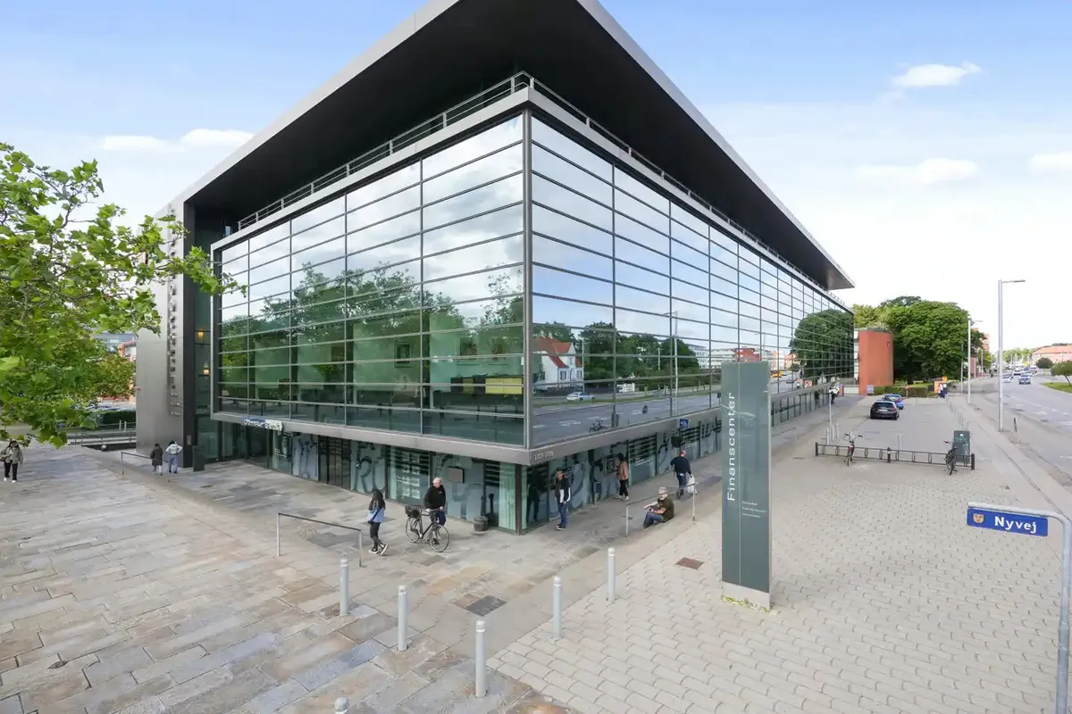 Glasfassade med refleksioner af bygninger og biler, flere mennesker går forbi, flere cykler er parkeret og gågade med