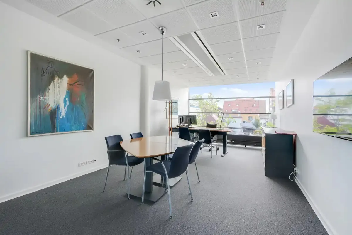 Kontor med bord, stole, skrivebord, computer, lampe og malerier på væggen med udsigt til byen fra vinduet