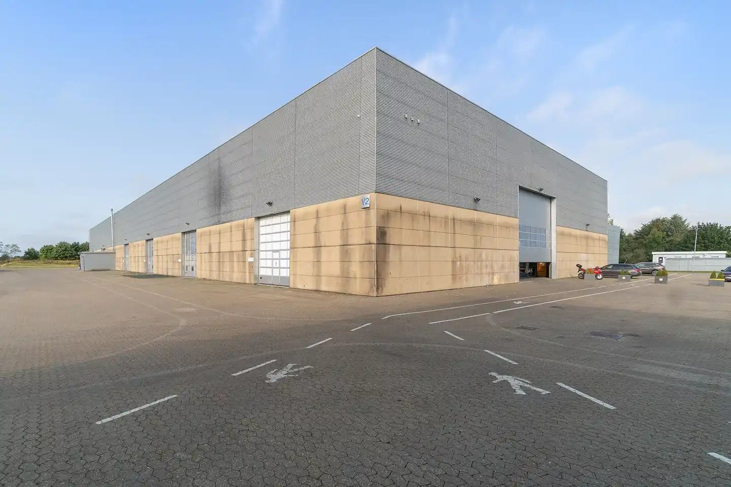 Et stort, enkeltværelses lagerbygning med nummer V2 i siden og store garagedøre. Bygningen har grå vægge og sandfarvede