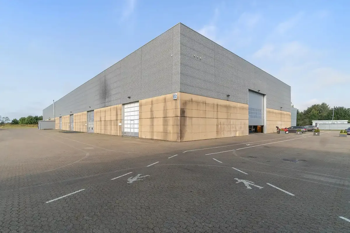 Et stort, enkeltværelses lagerbygning med nummer V2 i siden og store garagedøre. Bygningen har grå vægge og sandfarvede