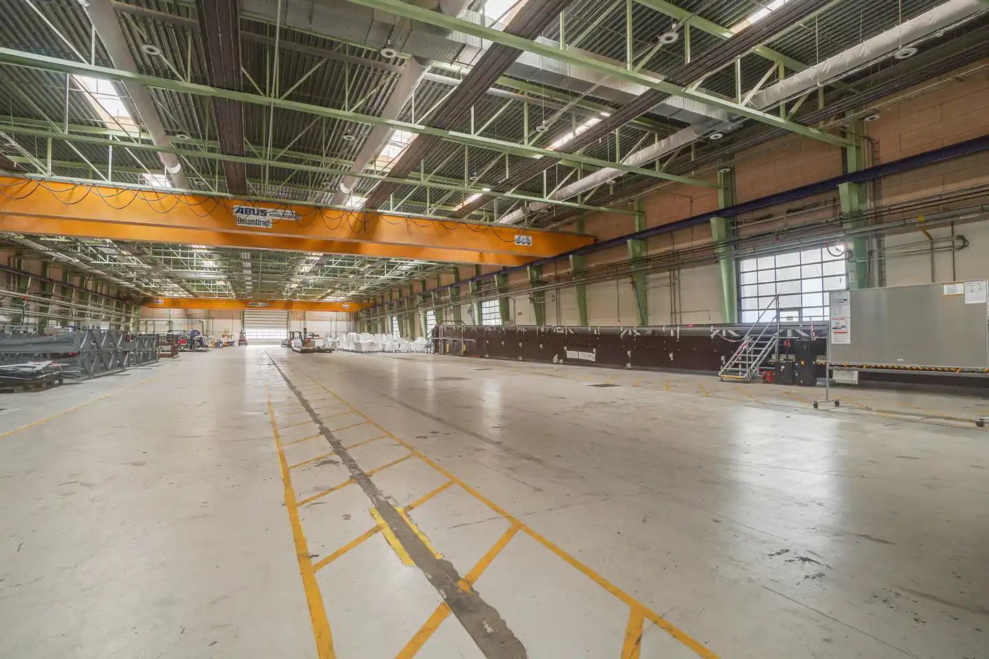 En stor, tom lagerhal med højloft og gule sikkerhedslinjer på gulvet. Et stort gult kransystem hænger fra loftet.