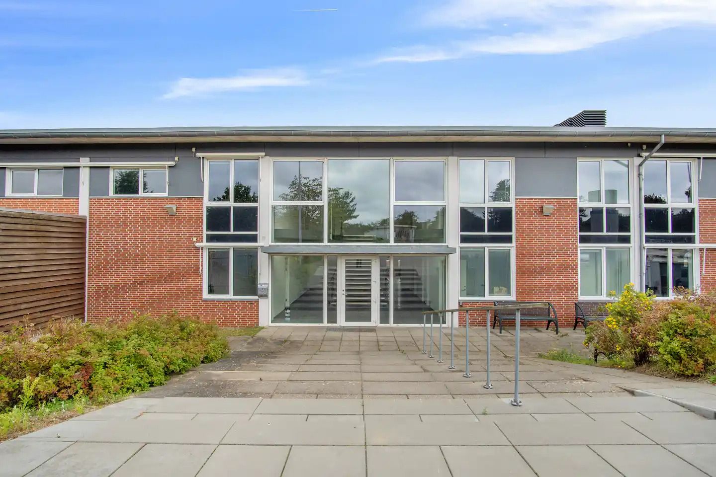 Kontorejendom med glasdøre og vinduer i rød mursten og lysegrå facade med to bænke og planter foran indgangen