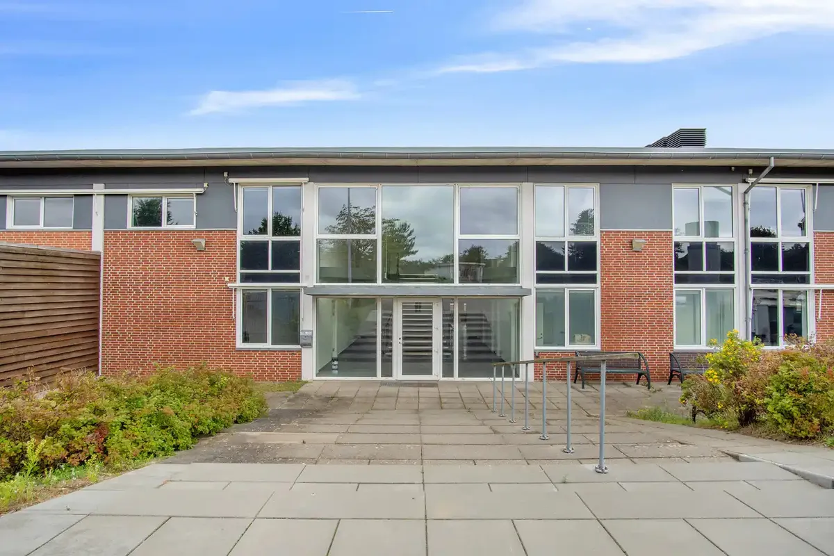 Kontorejendom med glasdøre og vinduer i rød mursten og lysegrå facade med to bænke og planter foran indgangen