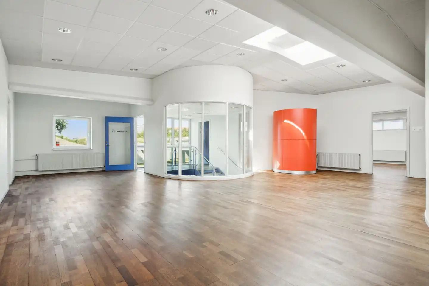 Kontorlokaler med højt til loftet og hvidt loft. Værelse med orange søjle og glasrute. Værelse med hvide vægge og blå dør.