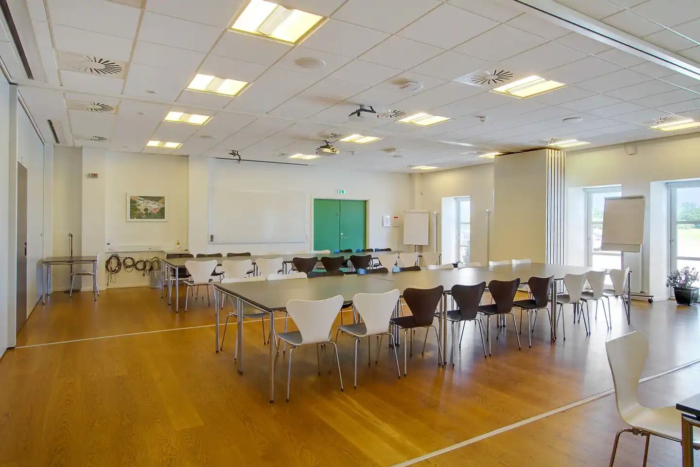 Moderne konferencesal med trægulve, lysekroner og store vinduer. Væggene er hvide med en tavle og et billede. Salen er tom