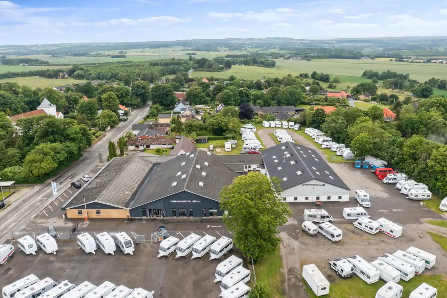 Mange hvide og røde campingvogne står parkeret i et stort areal med grønne træer og græs. Der er et stort og højt