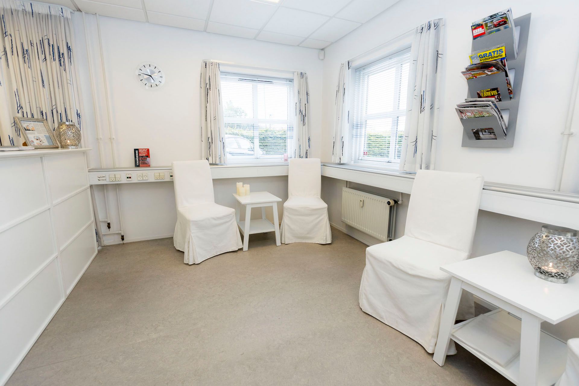 Hvid klinik med to stole og et bord, hvor der er en avisstativ og en plade med aviser
