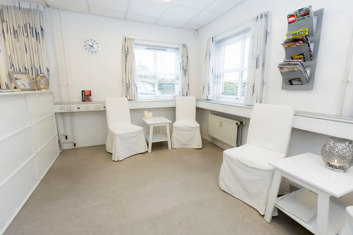 Hvid klinik med to stole og et bord, hvor der er en avisstativ og en plade med aviser