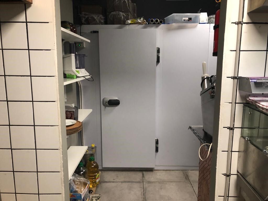 Hjørne af et køkken med køleskab, skabe, stående hylder og flere køkkenredskaber og apparater