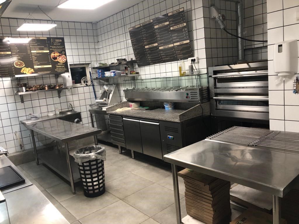 En ren og fuldt udstyret cateringskøkken med rustfri stålborde, en køleskab, pizzaovn, afløb, opvaskemaskine og flere