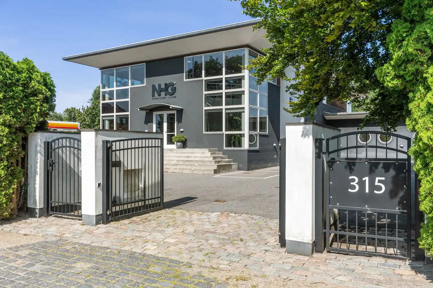 Moderne bygning med NRG-logo og nummer 315 på portene, omgiven af grønne planter og træer, og placeret i en velplejet og