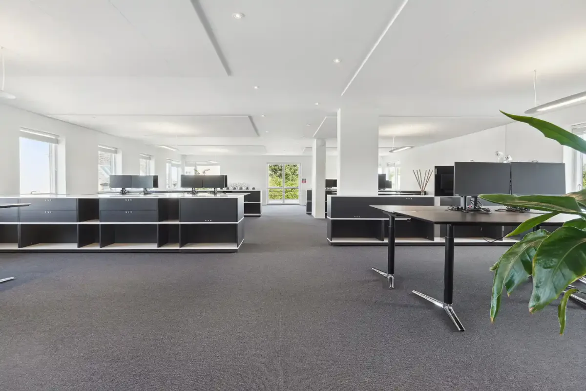 Moderne og minimalistisk indrettet kontor med højt loft, store vinduer og lysegulv. Bord og stole er sorte og hvide.